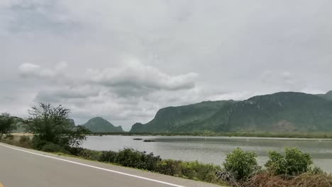 Fahren-Entlang-Einer-Straße-Neben-Einem-See-Mit-Bergigen-Hügeln-Im-Hintergrund-In-Thailand