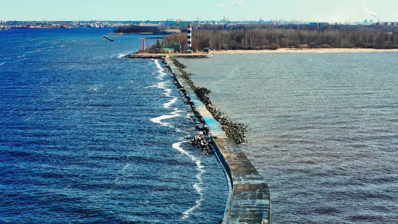 A long stone jetty cuts through bright blue Baltic Sea toward a tall black and white lighthouse dividing choppy waves from muted estuary waters and sandy coastline.
