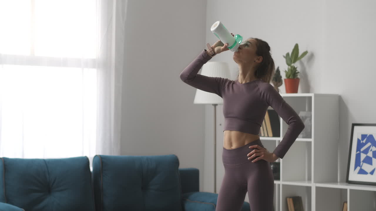 mujer adulta está haciendo fitness en casa bebiendo agua en el descanso de pie en la sala de estar estilo de vida saludable de la ama de casa moderna