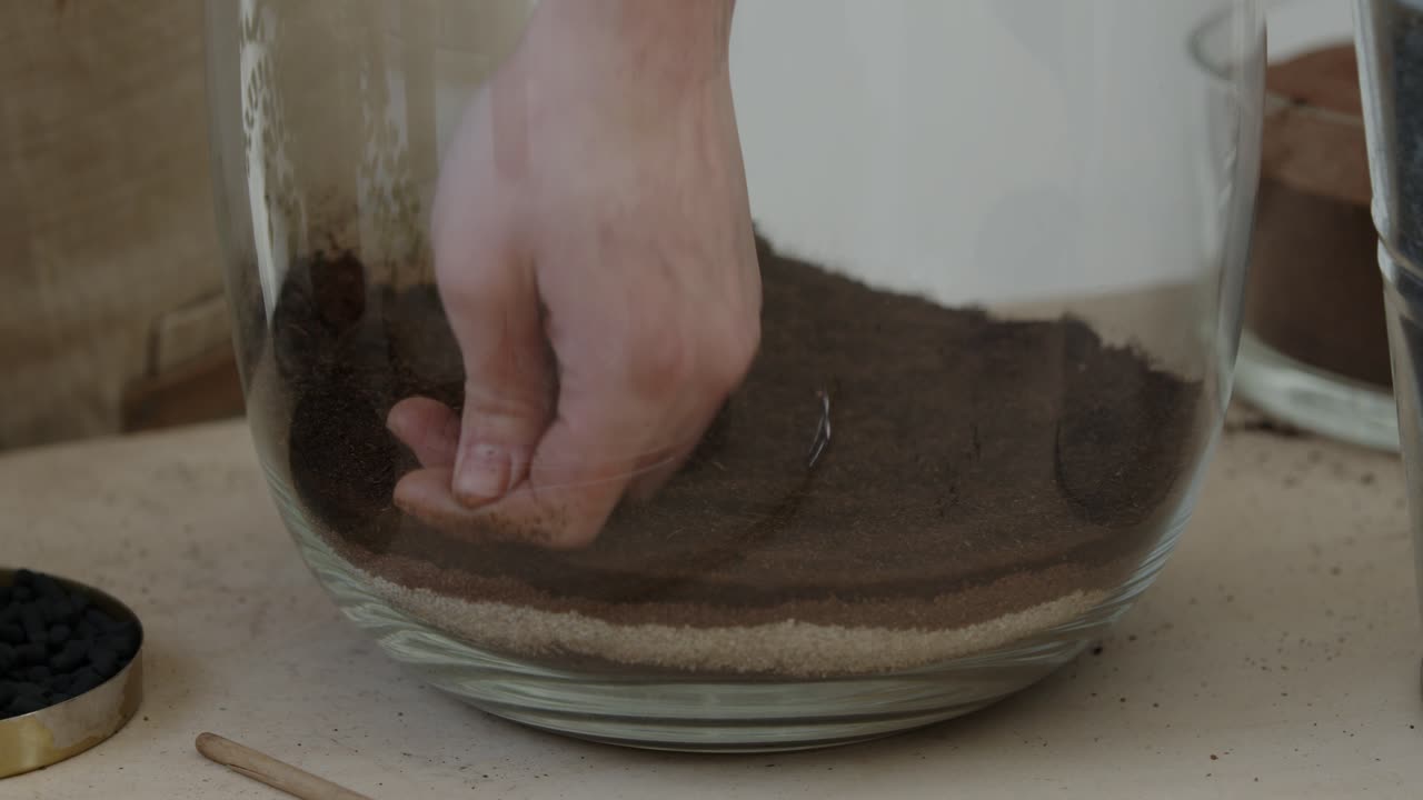 Una giovane botanica crea un piccolo ecosistema forestale vivente in un enorme vaso di vetro - preparando il terreno - un primo piano ravvicinato