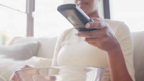 sección media de una mujer afroamericana sentada en el sofá, viendo televisión y comiendo palomitas de maíz, cámara lenta