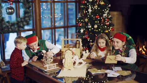 two elves are sitting at the table with a little boy and girl and show them letters of congratulations and christmas gifts on the background of the christmas tree and the fireplace near the window in the residence of santa claus