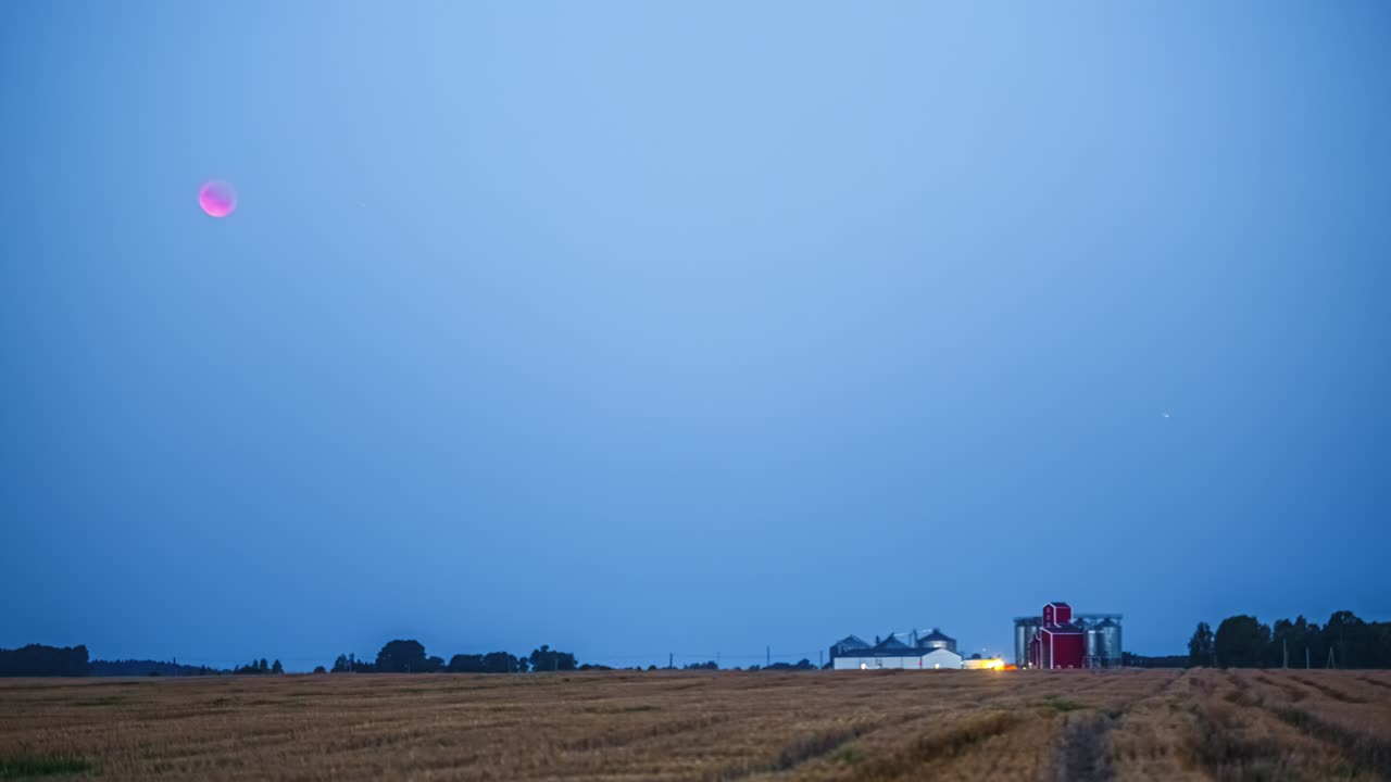 Warm red moon descends in warming glowing dark to light blue sky as farm lights turn on in morning