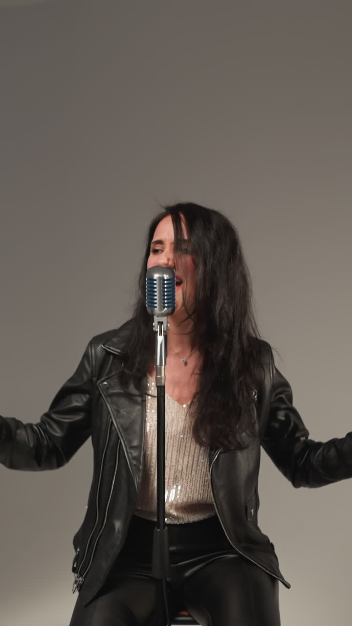 A vocalist in a black leather jacket sings passionately with expressive hands and eyes closed,in front of a vintage microphone.The camera zooms in and out, against a grey background