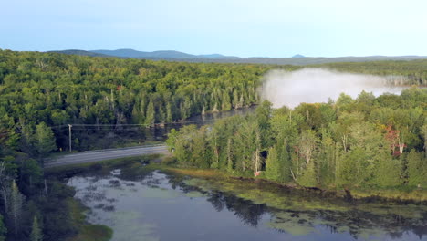 una toma aérea de un dron que emerge de la espesa niebla y la niebla para revelar las aguas azules del estanque de espectáculo y un camino que atraviesa el verde bosque del desierto de maine