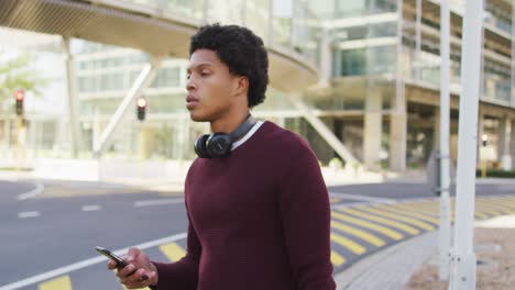 african american man in city, using smartphone, wearing headphones and backpack in street