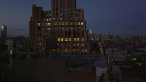 Forwards-fly-above-city-after-sunset.-Tall-office-building-with-lighted-windows-against-dark-sky.-Manhattan,-New-York-City,-USA