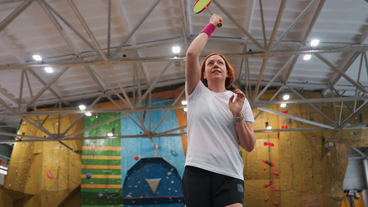 Caucasian woman playing badminton indoors focused training session in converted climbing gym, tossing shuttlecock, preparing high serve, executing upward swing, watching shuttle trajectory