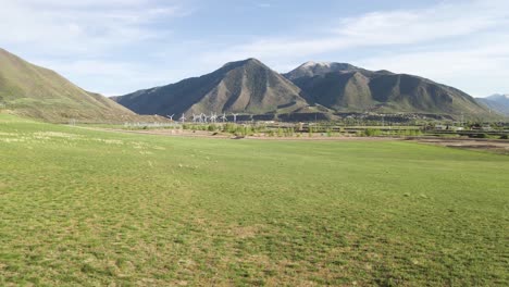 Sobrevuelo-Aéreo-Campos-Rurales-Verdes-Que-Muestran-Una-Granja-De-Turbinas-Eólicas-Ecológicas-Y-Montañas-Wasatch-En-Segundo-Plano.