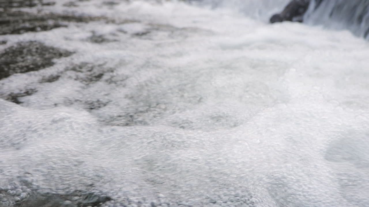 A detailed view of bubbling water in a flowing stream near a waterfall, captured in slow motion for a dynamic effect.