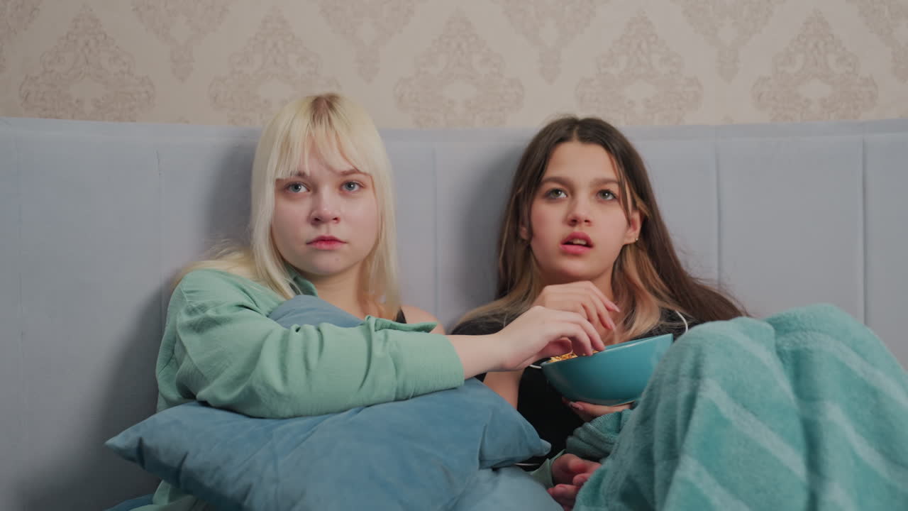 Two young girls sit closely on bed watching movie with serious expressions while holding pillows and eating popcorn, wrapped in soft blankets in cozy bedroom
