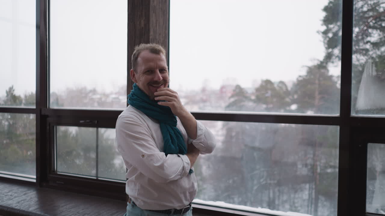 hombre se ríe contra la ventana panorámica. hombre maduro con pañuelo cambia la emoción de la reflexión a la sonrisa en la residencia. cambios de humor durante los días fríos