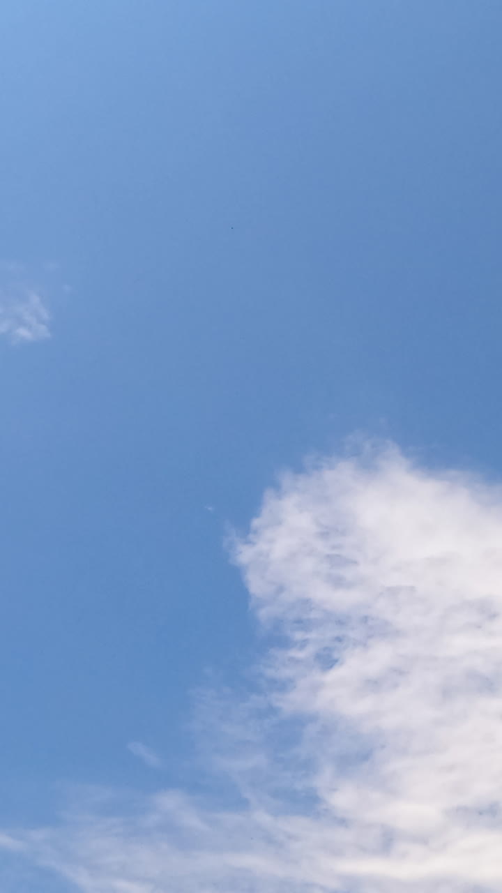 Blue summer sky with few light cirrus cloudscape floating by. Beautiful sunny day sky timelapse. Low angle view. Vertical video