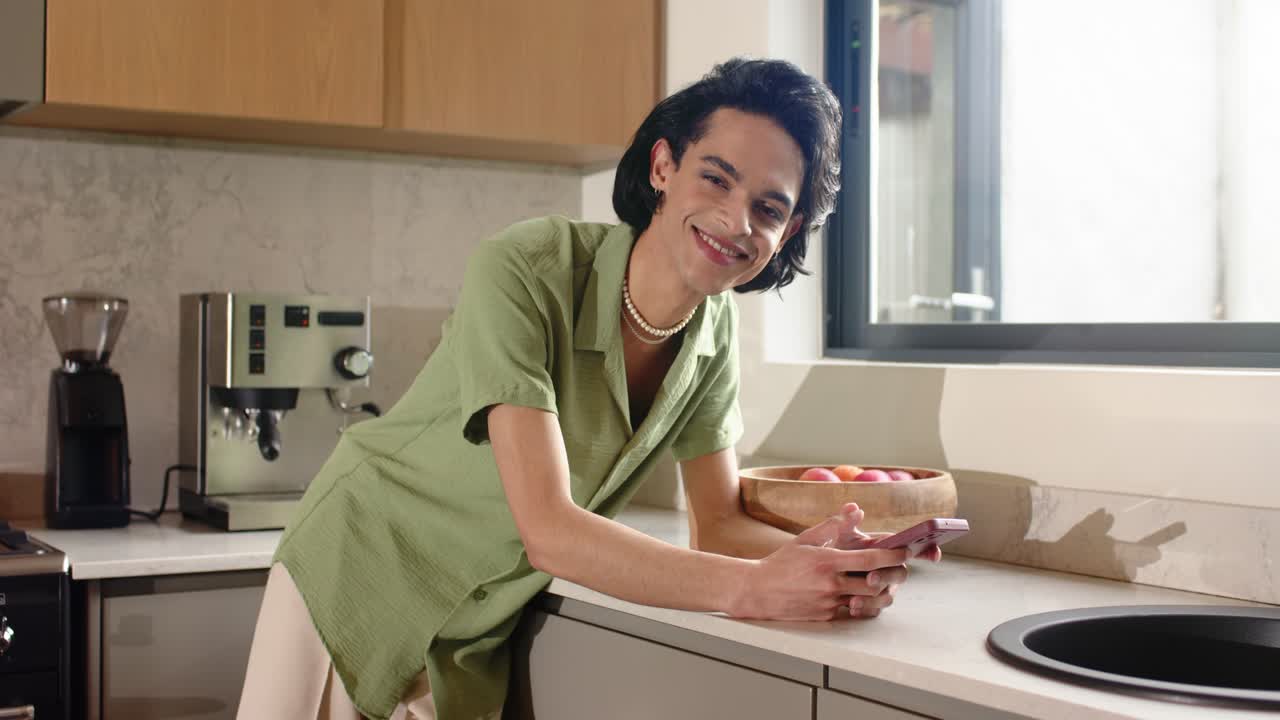 Uomo sui vent'anni che afferra lo smartphone dopo una notifica, scorrendo sul bancone della cucina accanto a una ciotola di frutta e sorridendo.