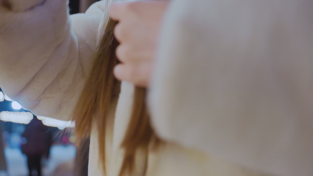 Close-up of an elegant woman in a cozy winter outfit combing her hair outdoors at night, warm glowing festive lights illuminate the scene, with a blurred background of people and cityscape