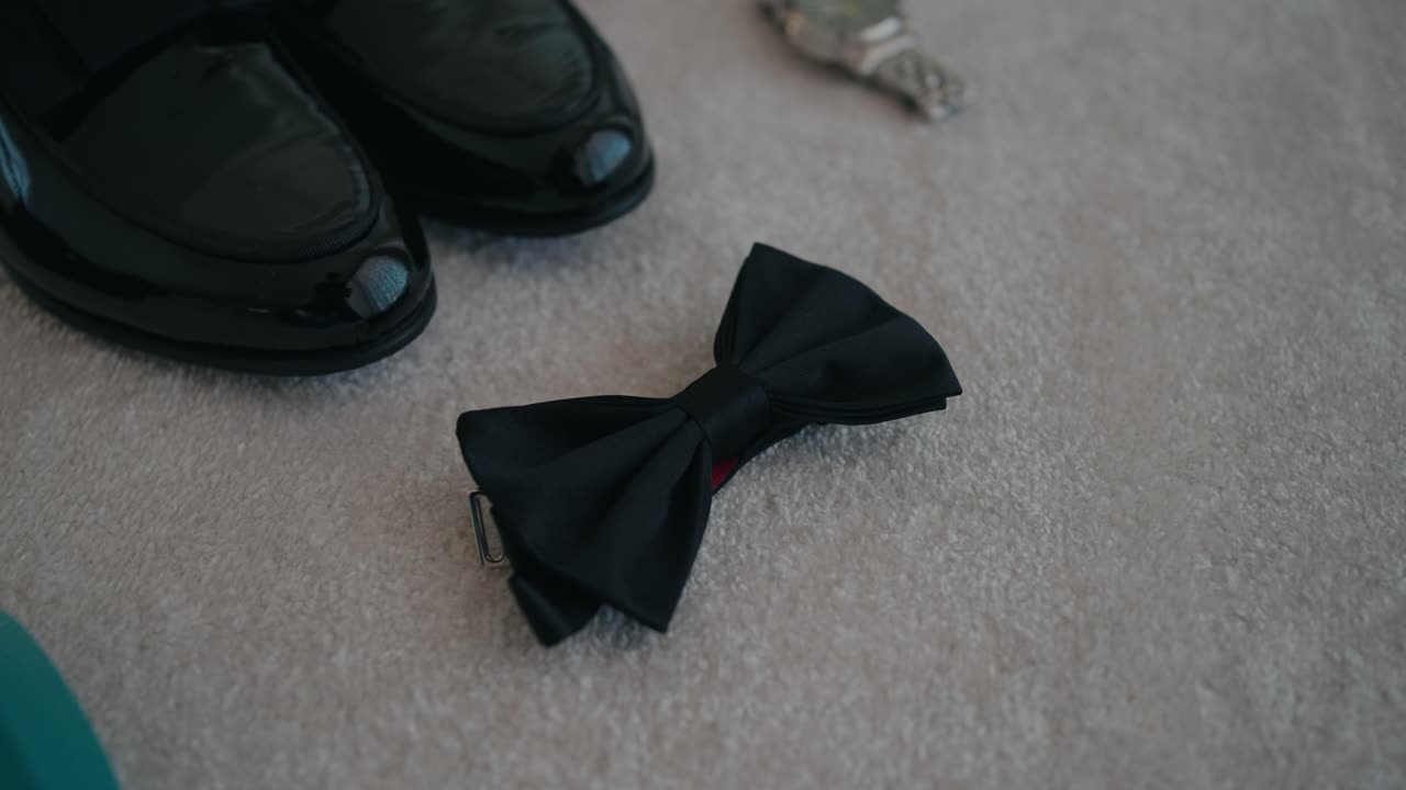 Close up of a black bow tie beside polished formal shoes on a light surface