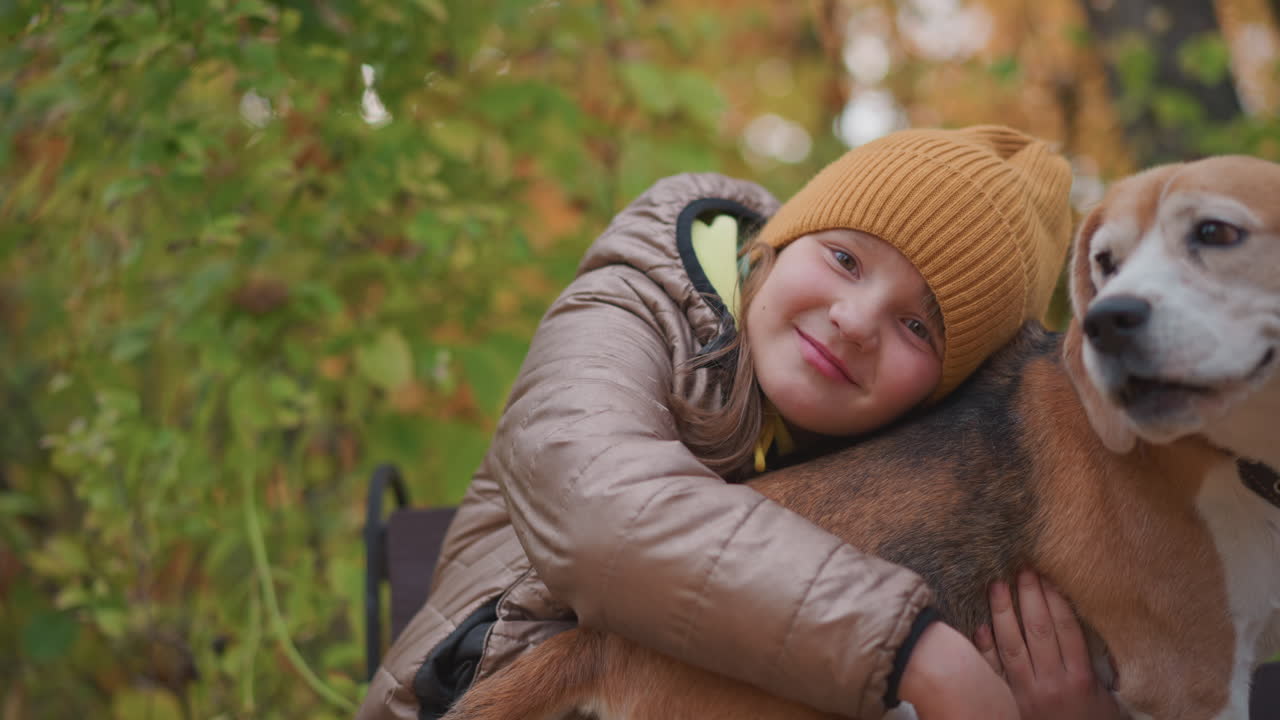 young girl in brown puffy jacket and mustard beanie gently rests cheek on loyal beagle's back, expressing warmth and affection amid colorful autumn foliage in quiet peaceful forest park setting
