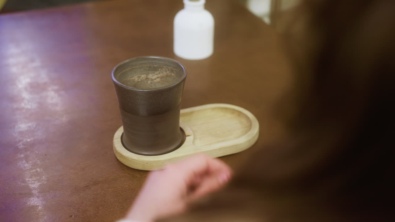 Close-up shot of woman's hand gently holding coffee cup on table, with soft lighting and cozy atmosphere, perfect for capturing moments of relaxation and enjoyment in a calm cafe setting