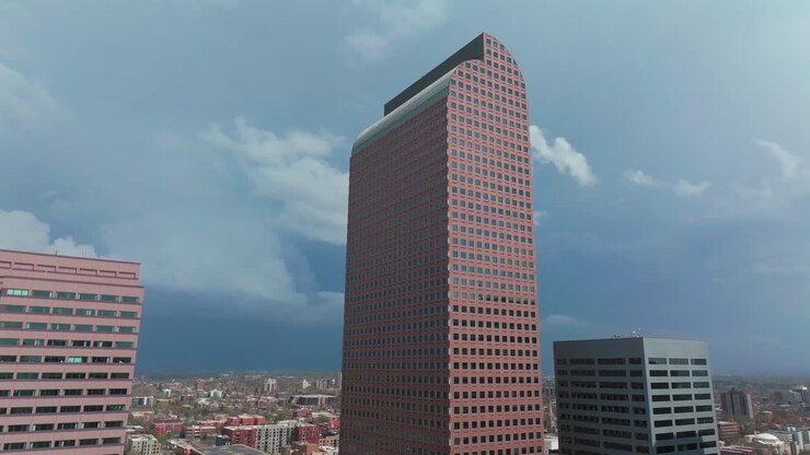 Paisagem urbana do centro de Denver, Colorado, com arranha-céus altos. Imagens aéreas de drone em uma tarde ensolarada, com sombra, sob céus azuis e nuvens de chuva tempestuosas. Destaques incluem Republic Plaza, City Park, Rhino, com movimento circular do drone para a esquerda.