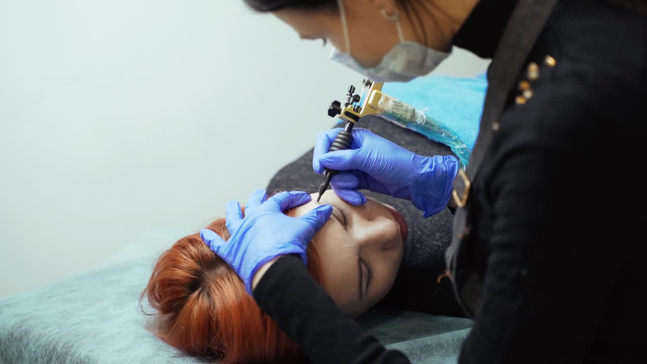 A woman in gloves is holding the client with one hand, and doing the eyebrow tattoo in the beauty salon with the other hand. Permanent makeup technique