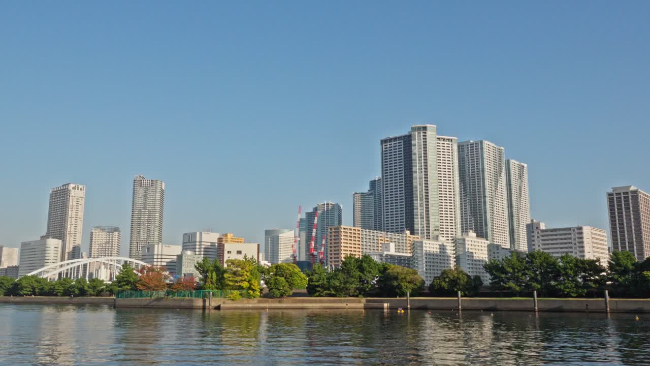 A wide view of Tokyo's modern skyline, featuring numerous high-rise buildings and a bridge, seen across a calm body of water.