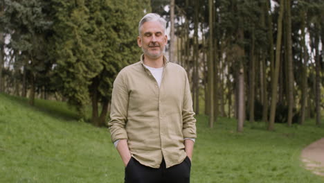 portrait of a gray haired man standing in the park with his hands in pockets and looking at the camera