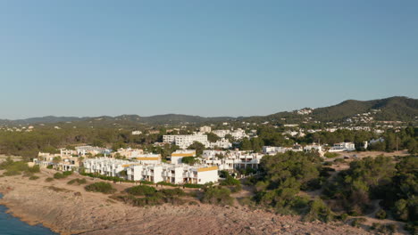 Aerial-forward-view-of-a-silent-and-calm-sea-surrounded-with-house-and-resort-villa-along-with-mountain-on-a-clear-sky-day-with-greenery-in-Ibiza-in-Spain