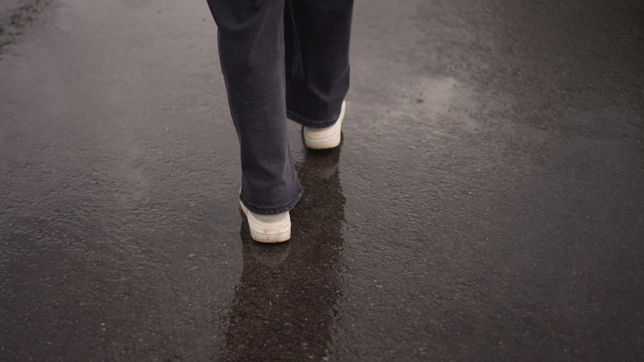 wet pavement closeup walking feet in sneakers, commuter strides through rainy urban street, reflective asphalt with puddles, dark jeans, slow steady pace conveying solitude and melancholy, overcast