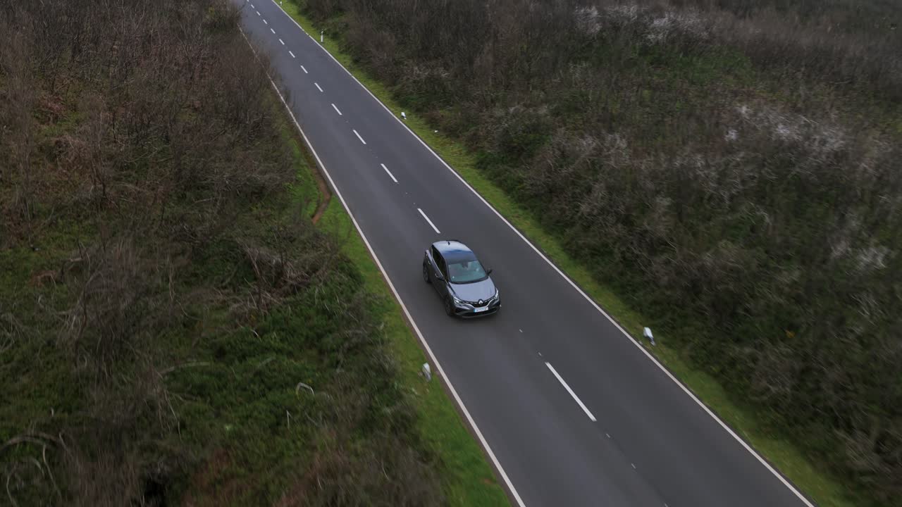 Aerial drone follows silver car driving along paved road through green and brown hilly, rural landscape in Porto Moniz region, Madeira, Portugal