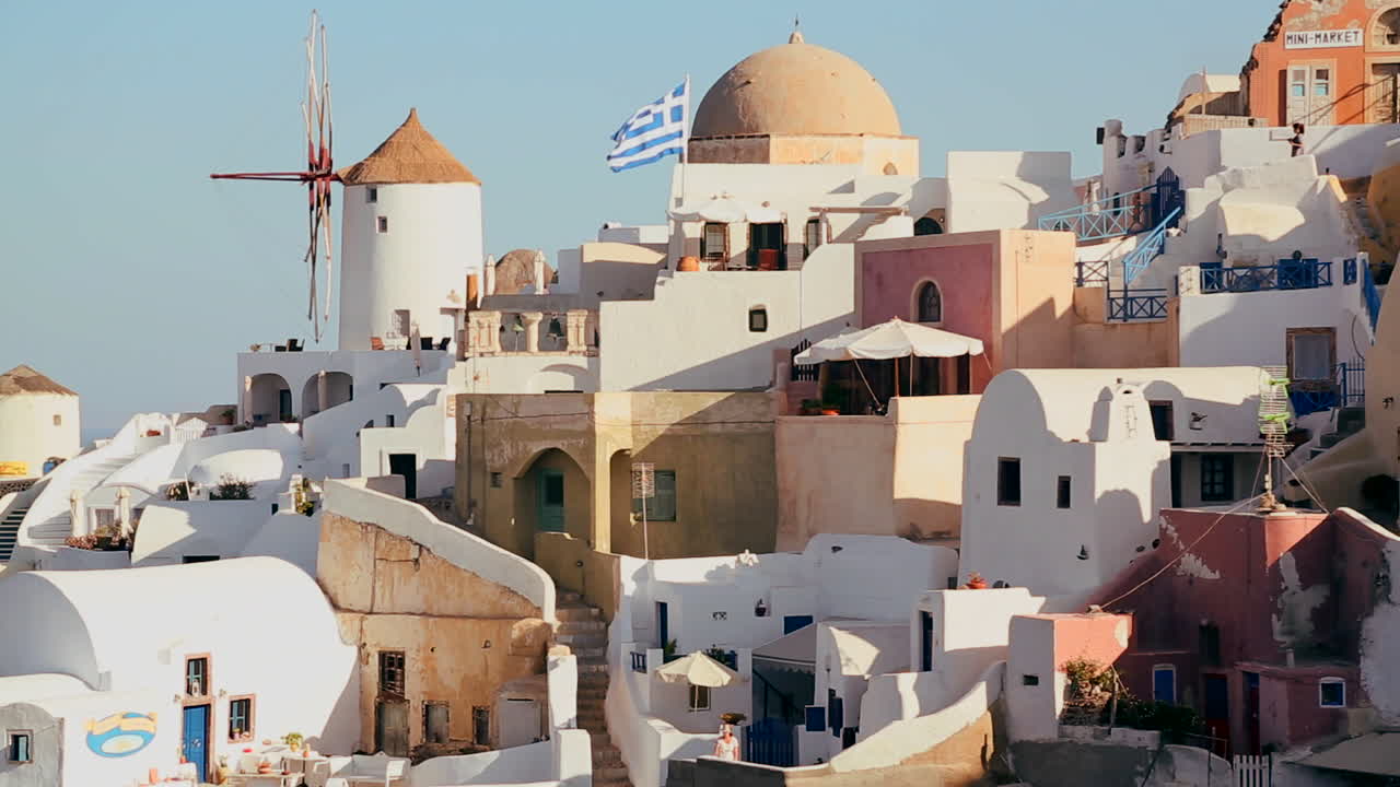 edificios blancos y molinos de viento bordean las laderas de la isla griega de santorini con una bandera griega en la distancia 1