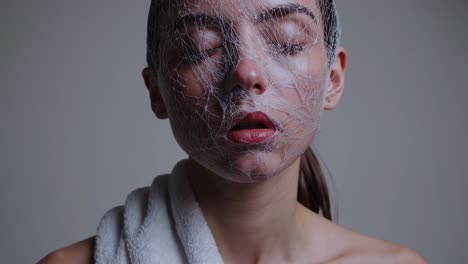 young woman wearing white mesh face mask, resting with closed eyes during beauty treatment, draped in white towel against neutral background