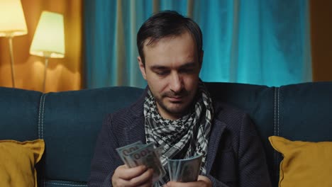 guy sitting on the couch by the table with a laptop and counting money for household expenses,