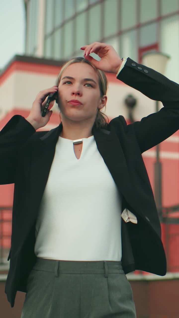 Frustrated white lady in professional outfit talking on phone looking tired and upset, walking near modern building with blurred crowd in distant background