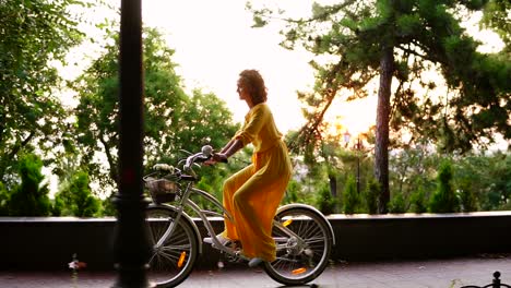 young brunette woman in long yellow dress cycling with flowers and packet in a basket and exploring the city. steadicam shot