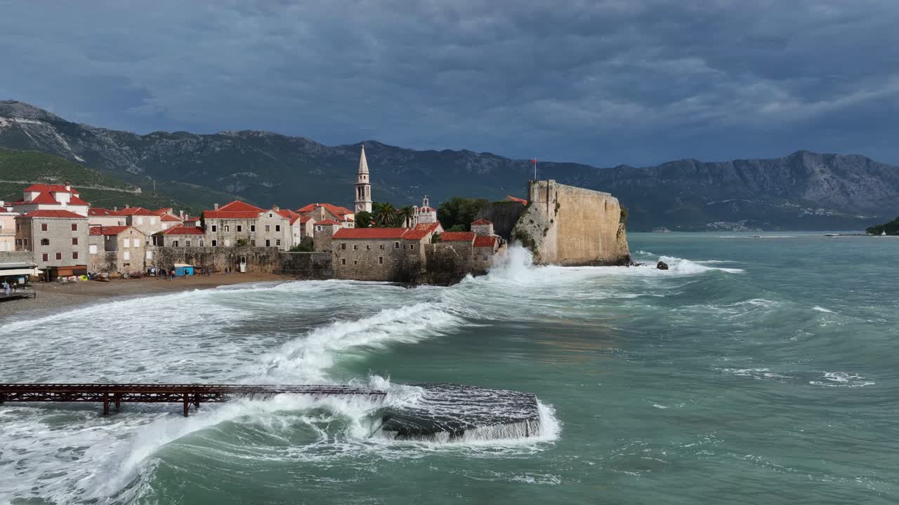 Aerial dolly parallel to Budva coastline with storm clouds looming over turbulent Adriatic waters as waves crash on dock