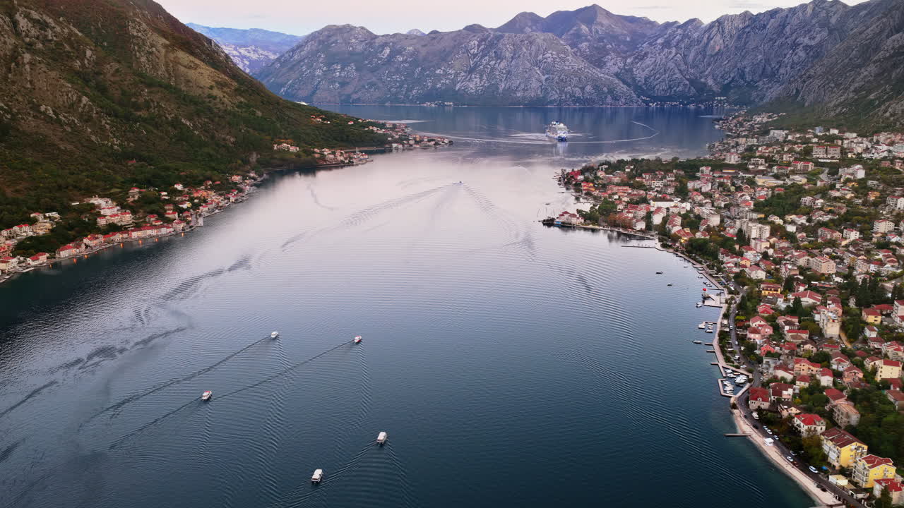 Aerial drone view of the Bay of Kotor surrounded by rugged mountains, with a cruise ship sailing in the center and coastal villages on both sides
