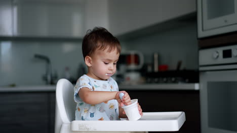 little kid eating yogurt from a plastic cup. toddler boy uses a spoon and fills it himself. blurred backdrop.