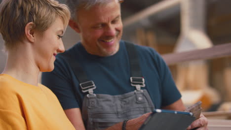 female apprentice learning from mature male carpenter with digital tablet in furniture workshop