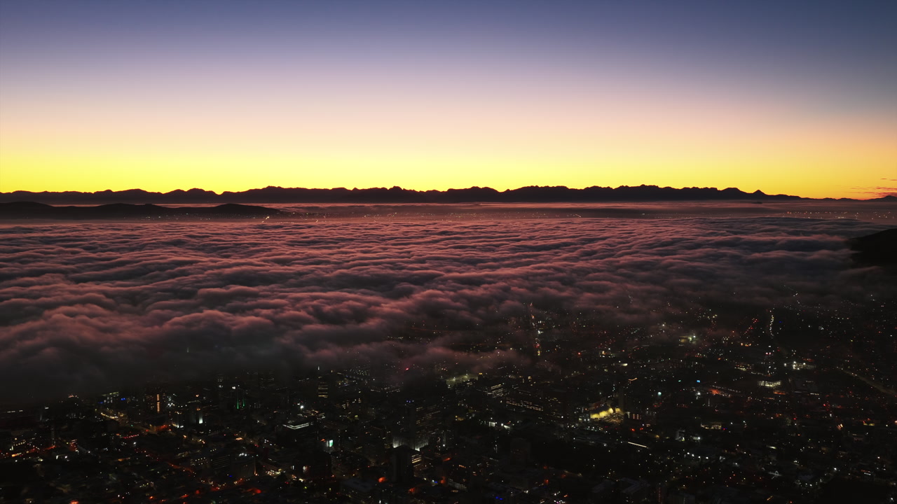 An aerial drone view captures Cape Town waking beneath a glowing sunrise, with thick clouds blanketing the city while golden light paints the horizon.