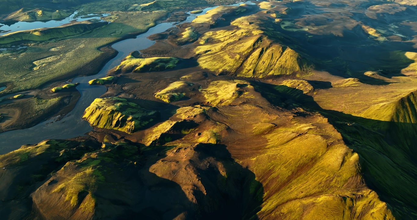 Fast drone flight over Iceland highlands volcanic ridges with mossy slopes, dark lava valleys and a winding river, golden-hour light creating dramatic shadows.