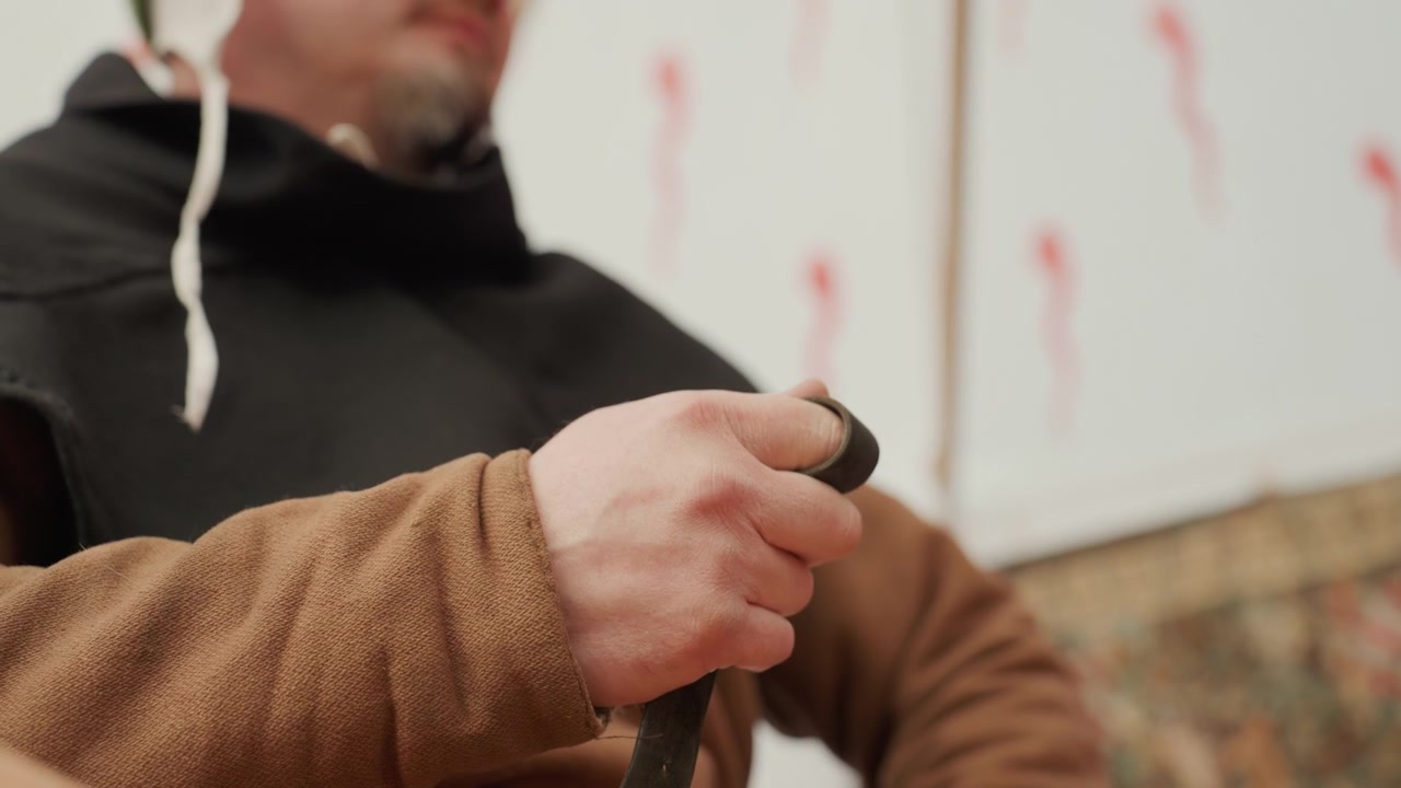Close-up van zittend persoon die met beide handen een donkere riem over de borst trekt en vastmaakt, met zichtbare vingerbeweging, stevige grip, casual kleding, warm binnenlicht en wazige achtergrond