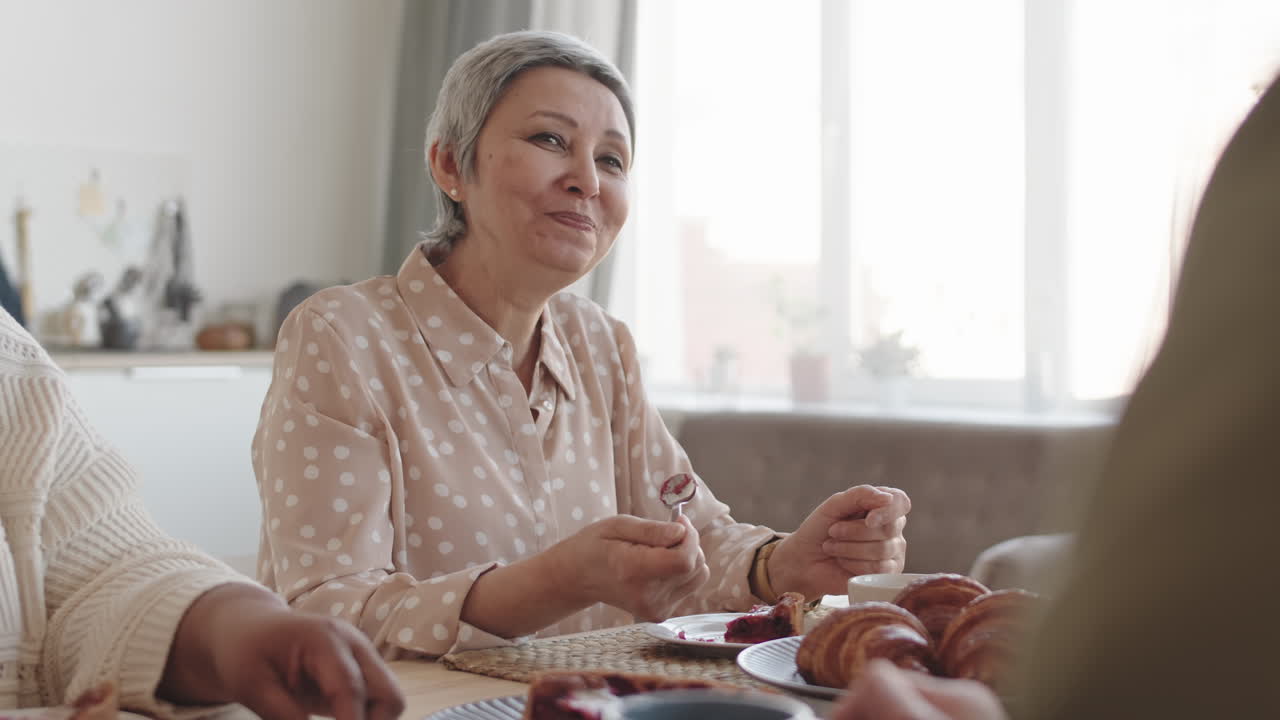 Tilting up of joyful Asian senior woman smiling, eating delicious sweet pie, drinking tea from cup, sitting at table, talking to unrecognizable company