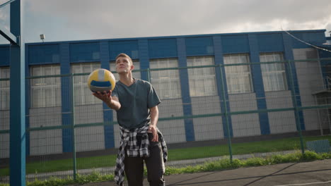 young athlete rotating volleyball in hand before throwing it up and serving it near basketball court, with blue building and greenery in background