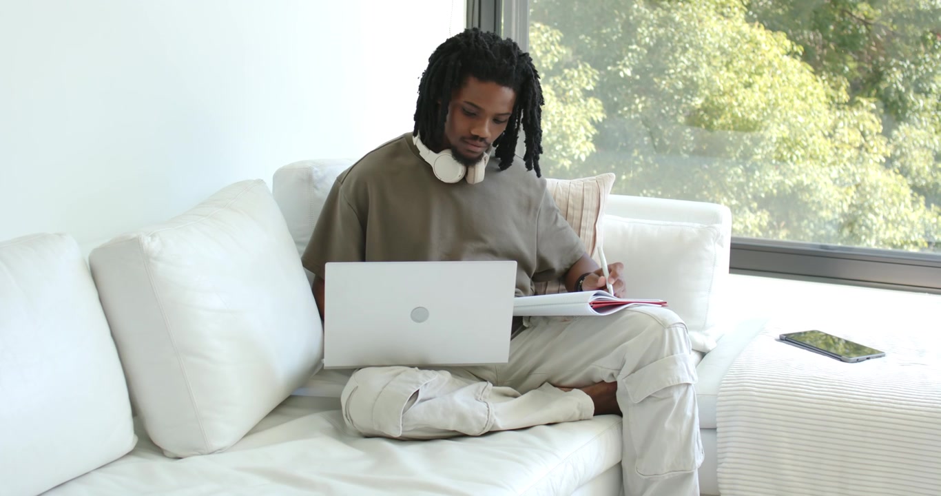 Man sitting on sofa at home with headphones, checking laptop and writing in notebook for work