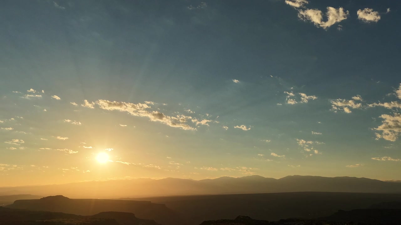 A 4K drone timelapse showcasing a striking blue and gold morning sky. Warm rays of sunlight drift smoothly across a sweeping high desert landscape, illuminating distant mountain ridges at dawn.