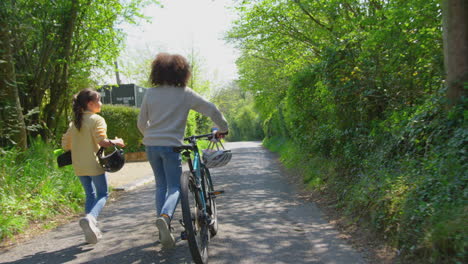 Vista-Trasera-De-Un-Niño-Con-Bicicleta-Y-Una-Niña-Con-Patineta-Caminando-Juntos-Por-Un-Camino-Rural