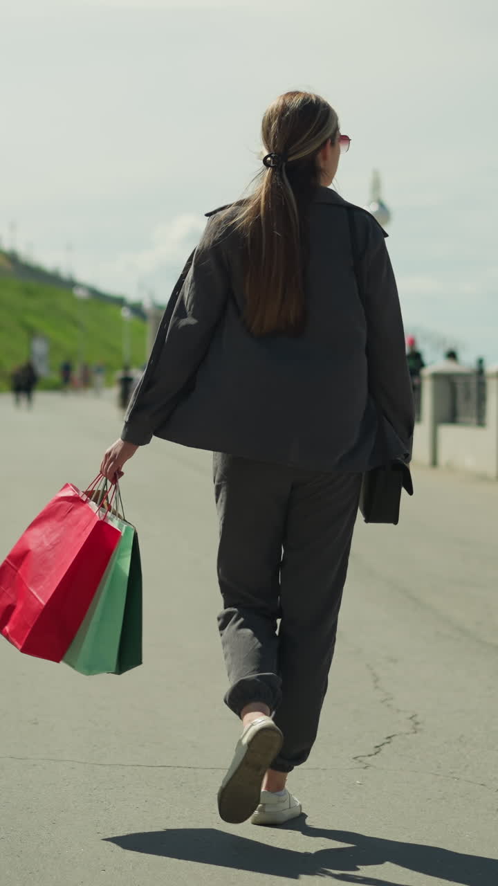 회색 옷을 입은 여인이 빨간색, 민트색, 녹색 쇼핑백을 들고 한 쪽에는 전등 기둥이 있고 다른 쪽에는 언덕이 있는 다리 위를 고 있다.