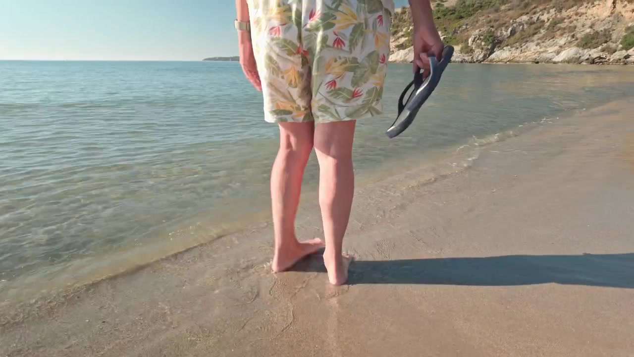 Legesyg kvinde køler fødder i Middelhavets bølger ubekymret på en fredelig strand. Panorer op