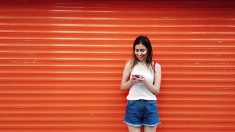 Una-Joven-Hermosa-Usa-Un-Smartphone-Frente-A-Un-Fondo-Naranja-Y-Rojo