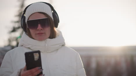 elegant young girl wearing oversized sun glasses and headset spins her phone around body while standing in snowy park at sunset, white coat and beanie glowing in soft light as hair flutters in breeze